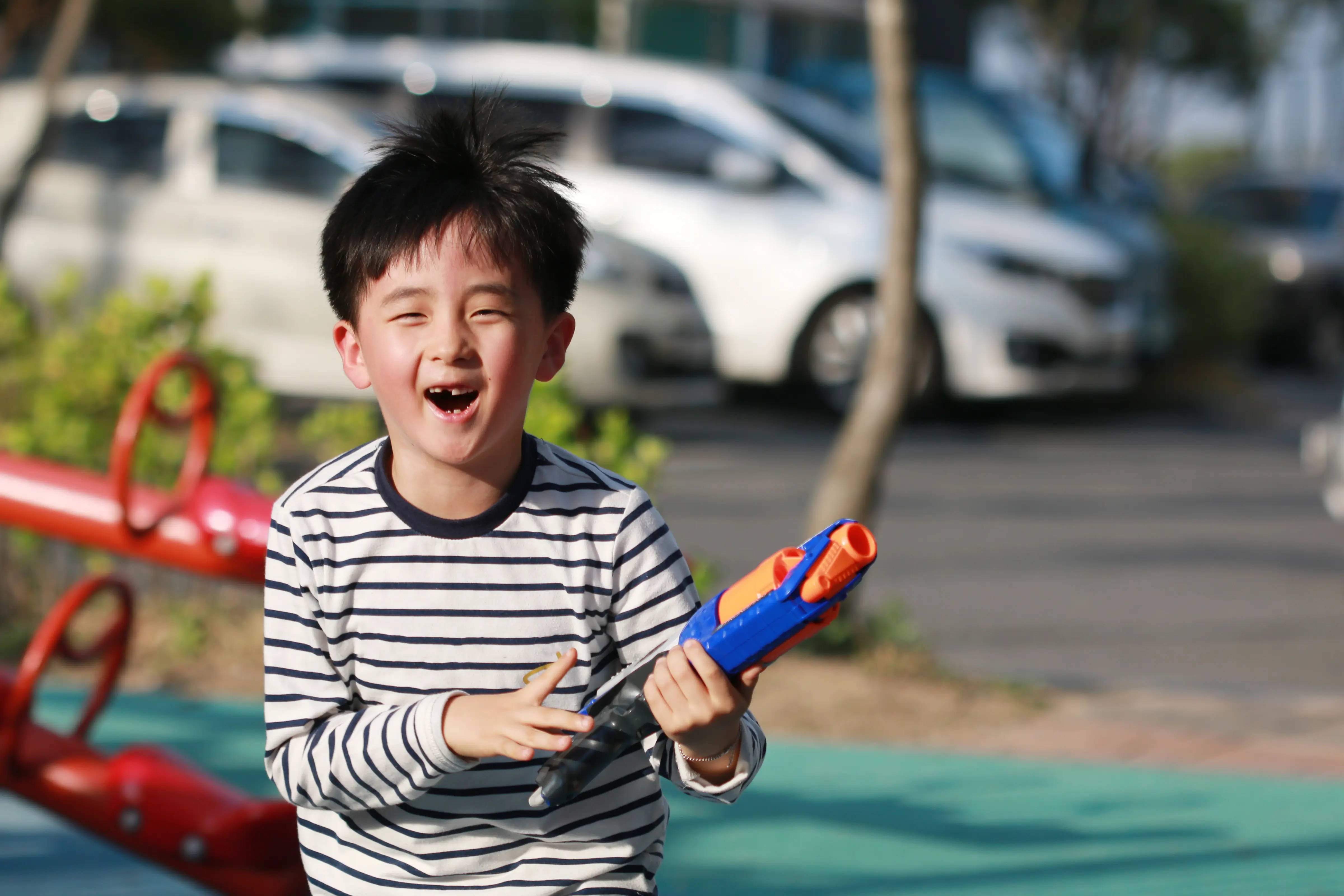 Young boy with missing tooth wearing white top with black stripes, smiling and holding a blue and orange Nerf gun