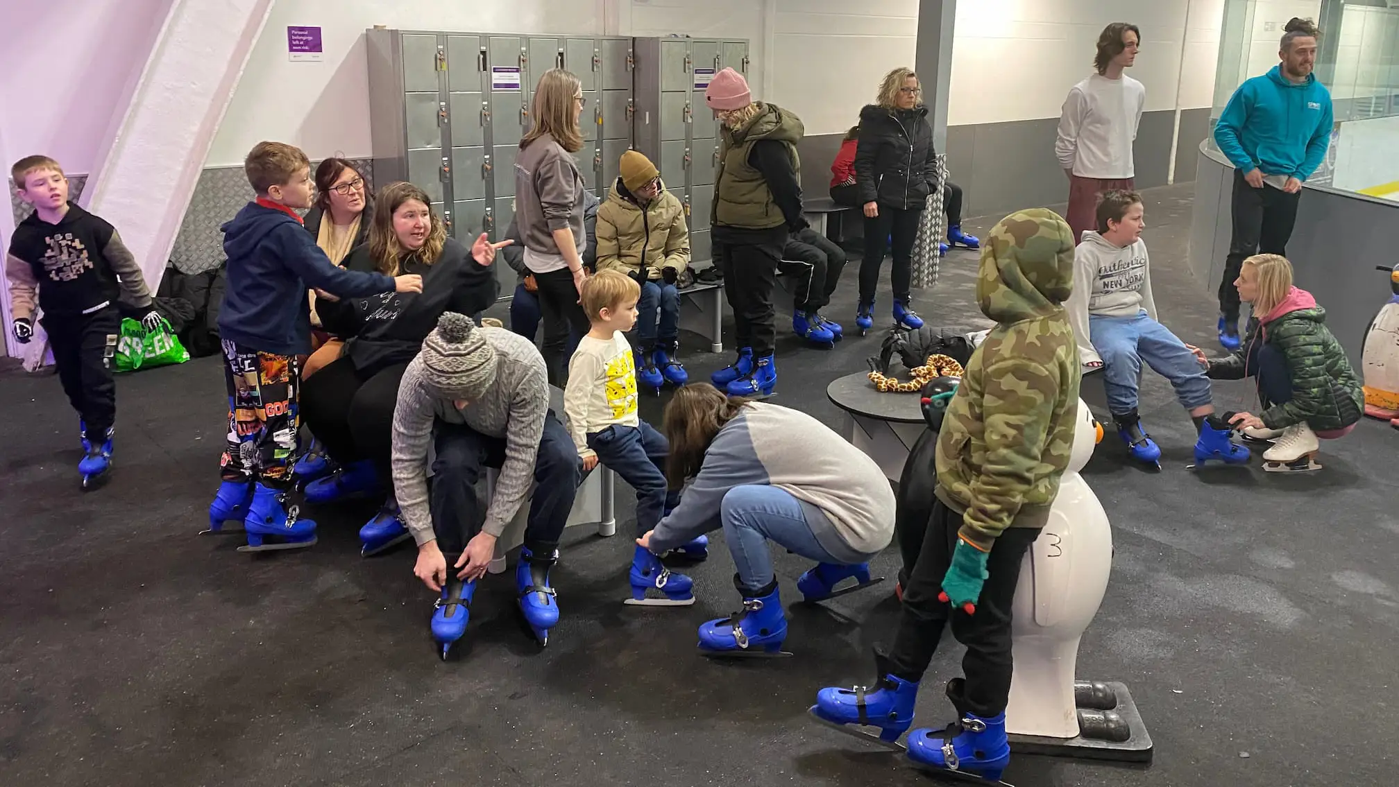 Group of adults and children sat on benches putting on blue ice skates at an indoor ice rink