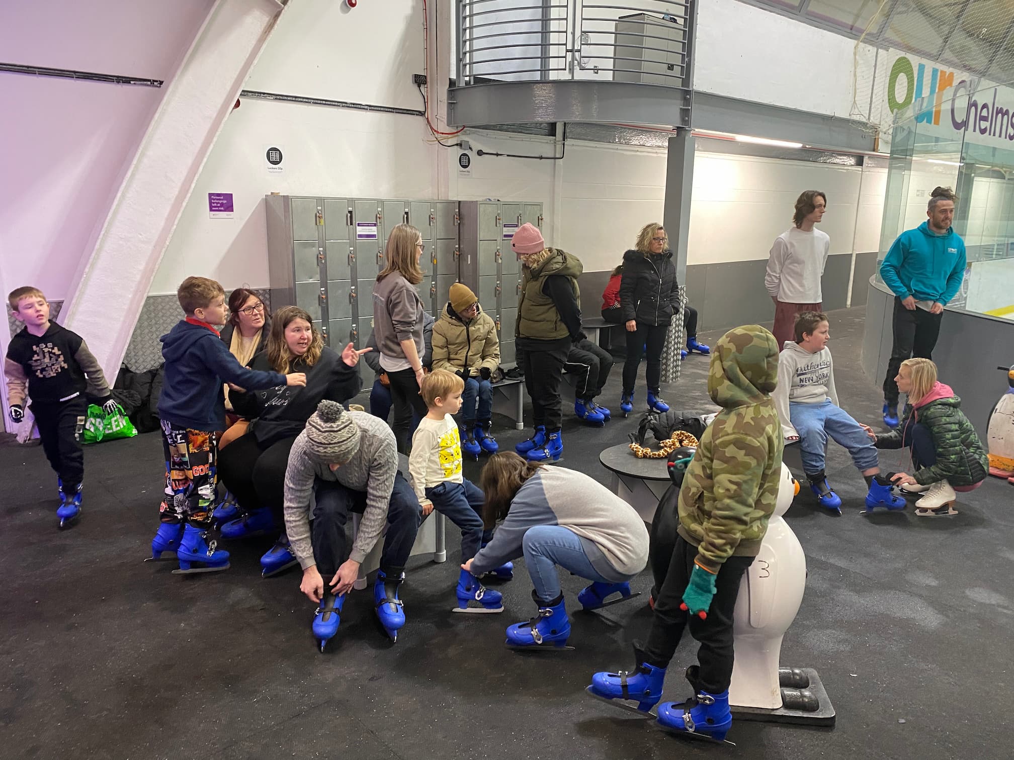 Group of adults and children sat on benches putting on blue ice skates at an indoor ice rink preview