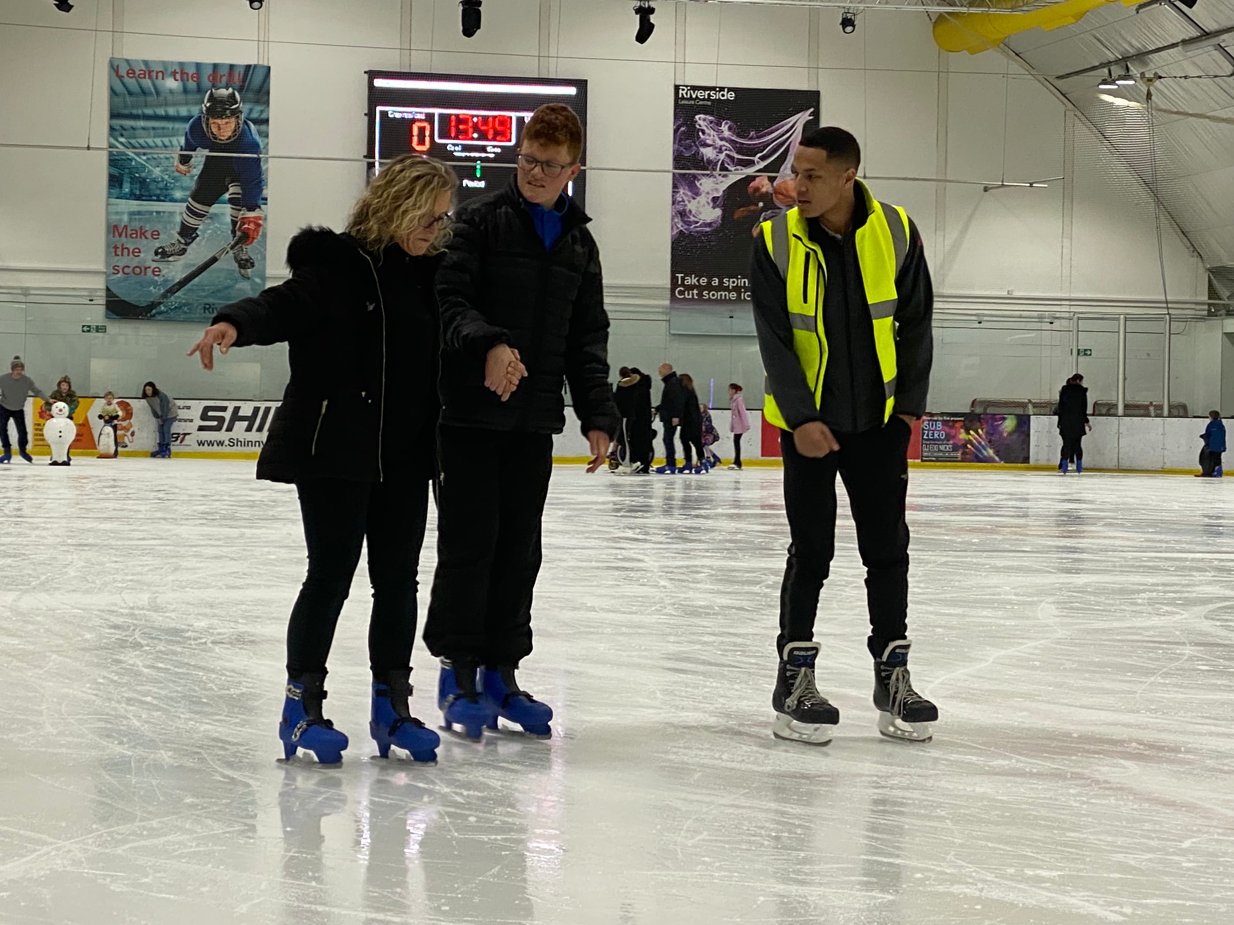 Two people learning to skate, being taught by a man in a high visibility jacket preview