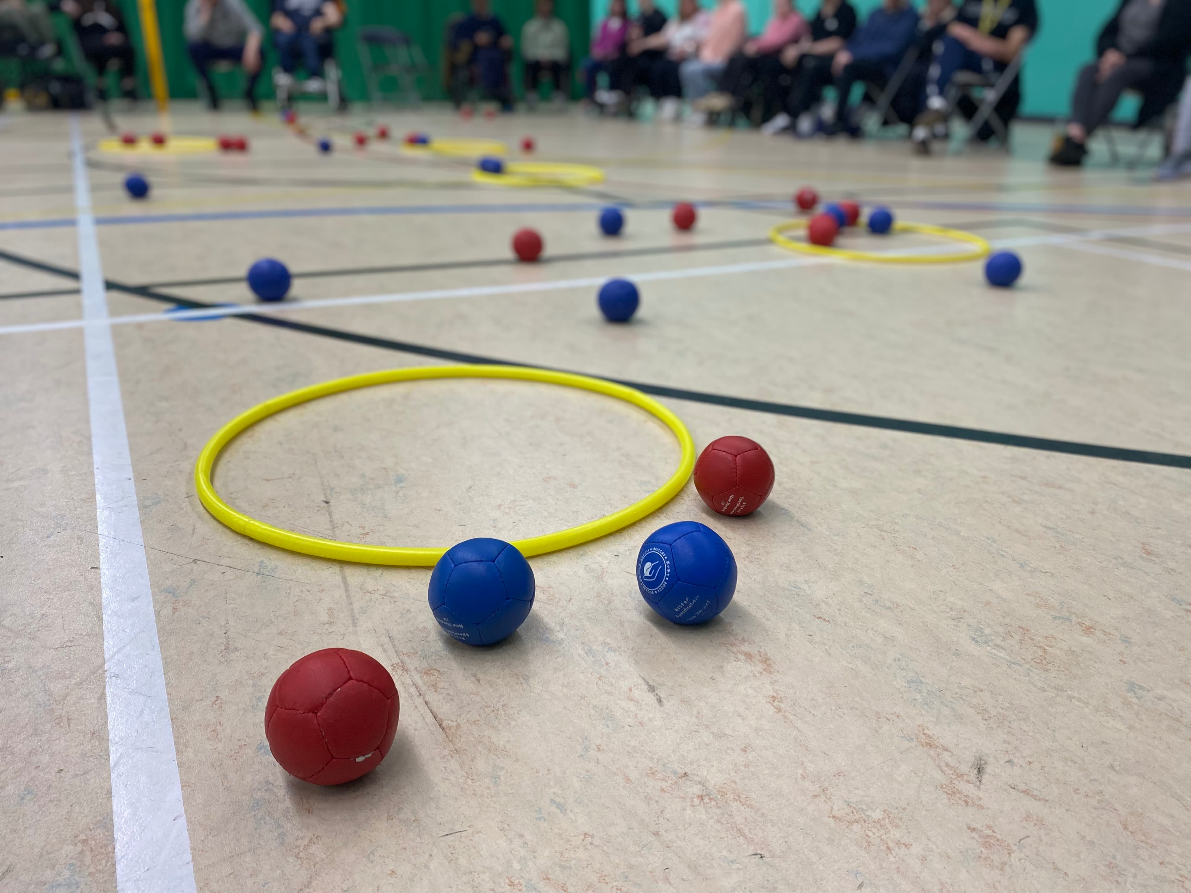 red and blue Boccia balls on the floor of a sports hall, next to a yellow hoop preview