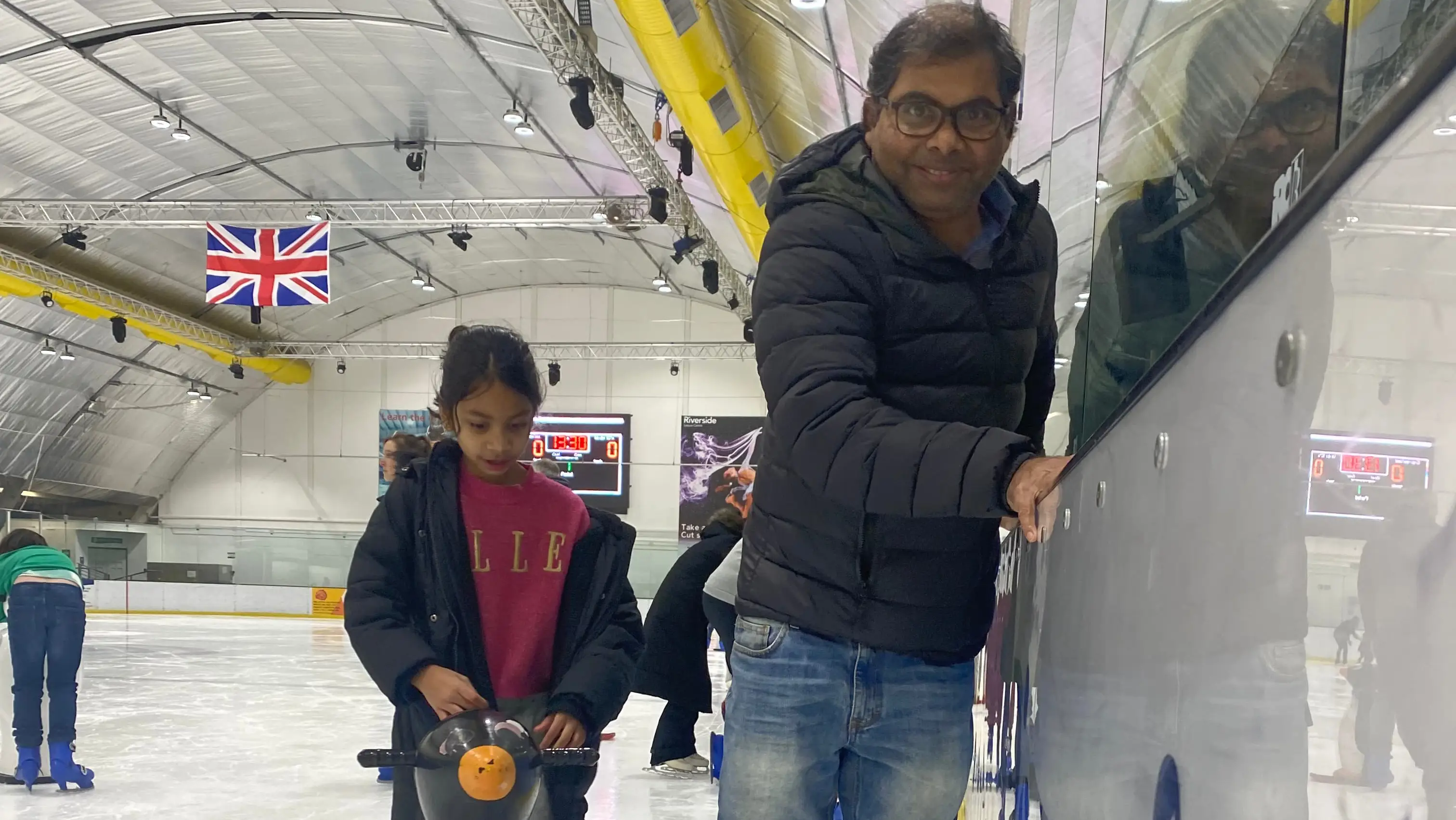 Father and daughter learning to ice skate, smiling.