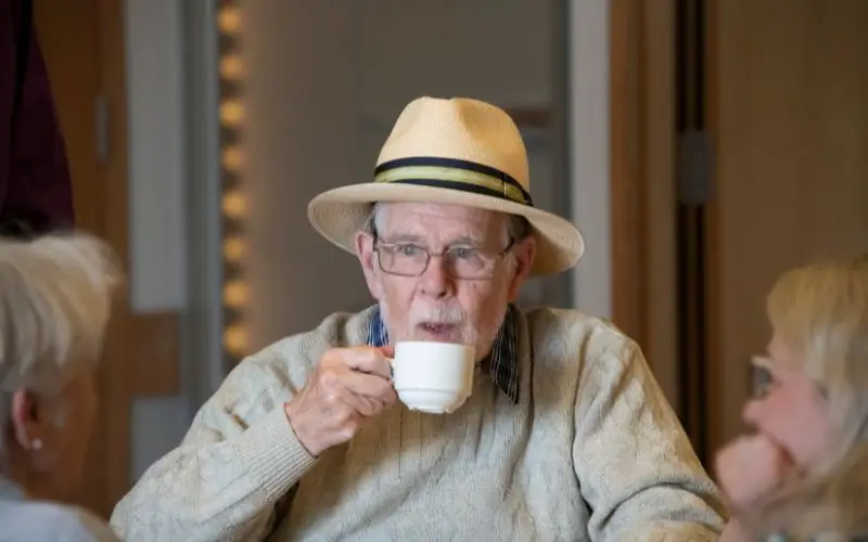 A man sipping a cup of tea while sat around a table with others. 