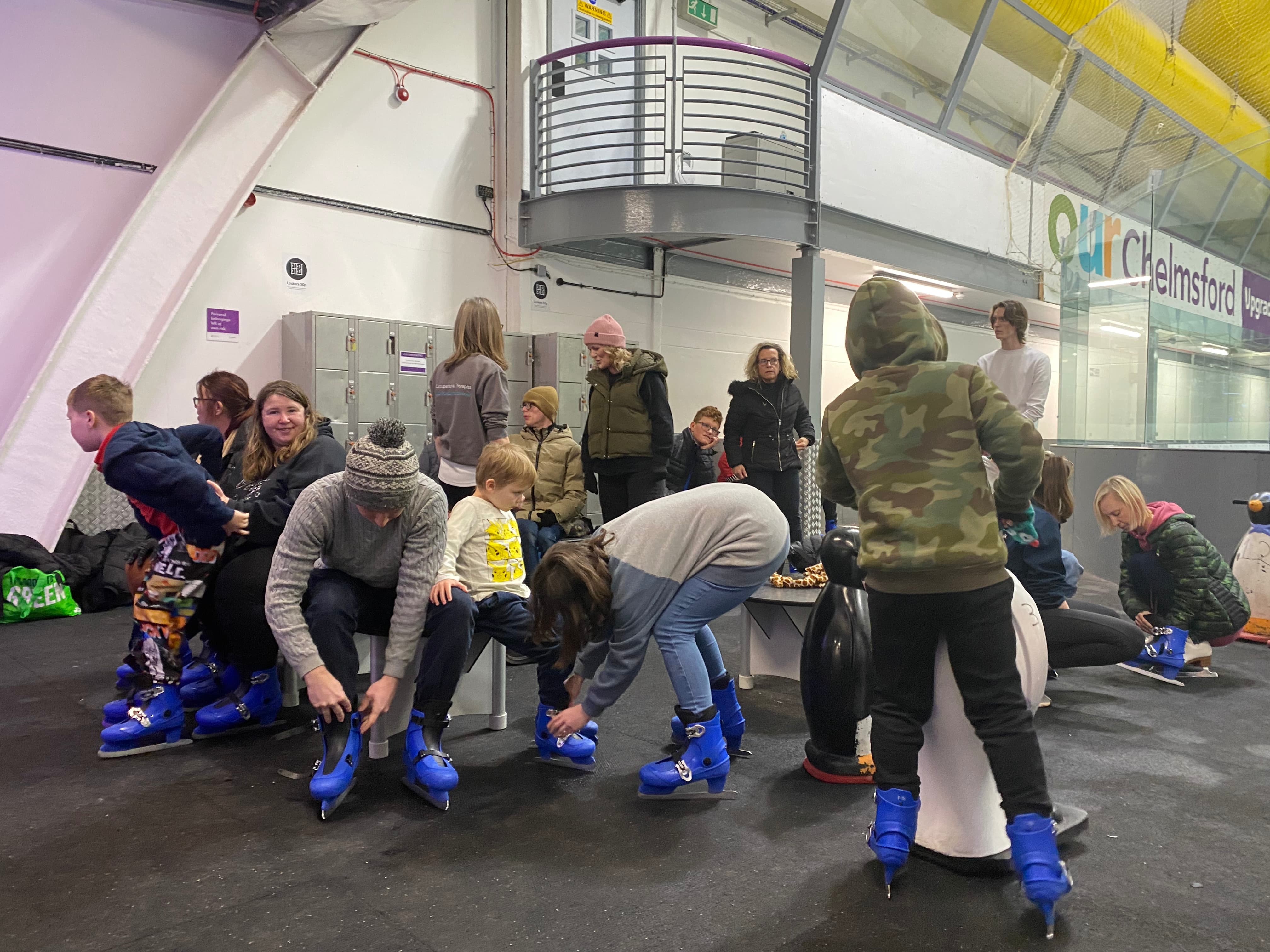 Group of adults and children sat on benches putting on blue ice skates at an indoor ice rink preview