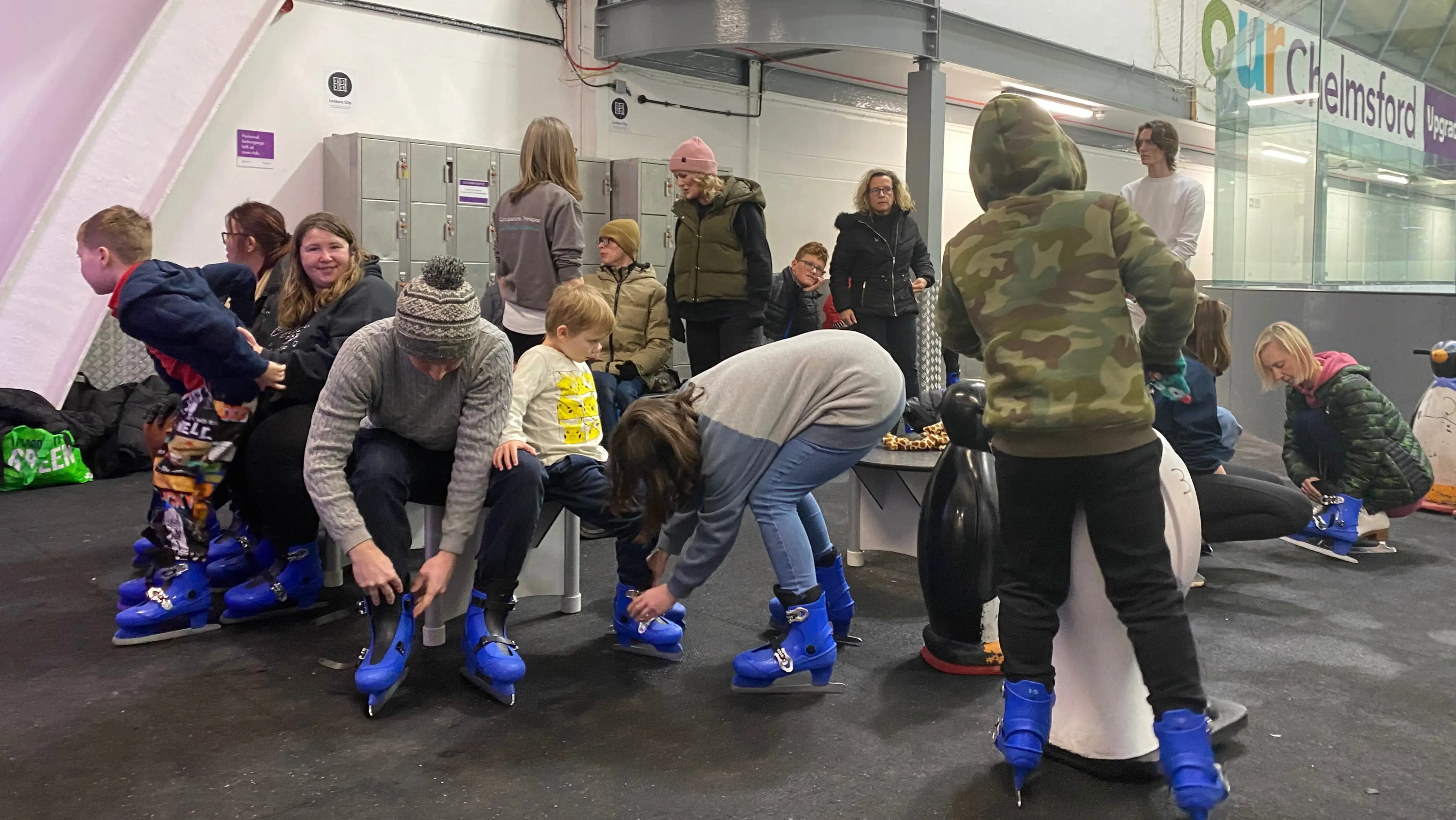Group of adults and children sat on benches putting on blue ice skates at an indoor ice rink