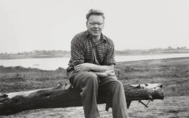 A black and white photo of JA Baker sitting on a log by the waterside. 