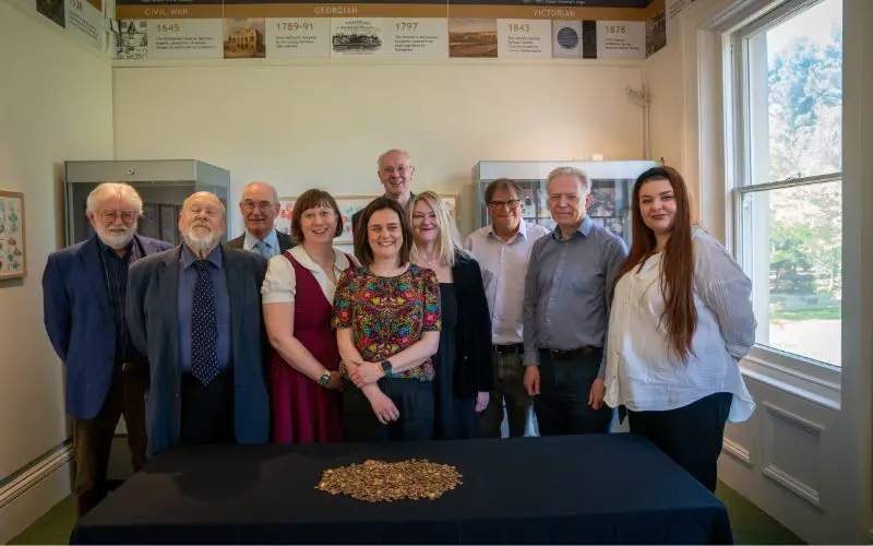 A group of people standing behind a pile of gold coins on a table. 