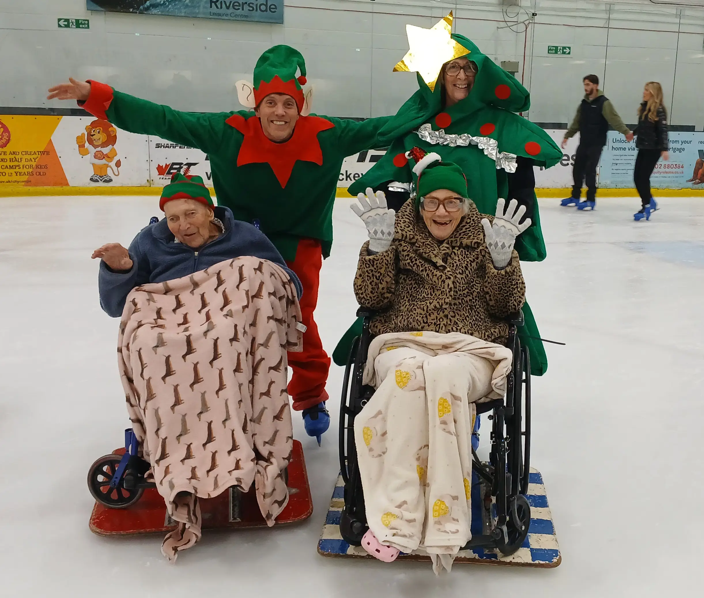 Two older people in wheelchairs with carers using boards on ice rink