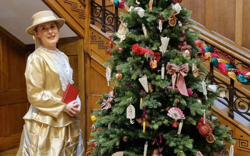 Rachel wearing a long gold dress and standing beside a Christmas tree decorated with Victorian style ornaments. 